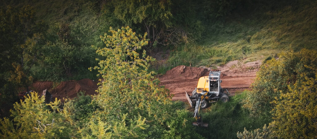 Drohnenaufnahme eines Baggers beim Wegebau entlang eines Waldwegs.
